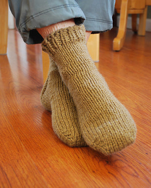 Legs from the midcalf down, feet crossed, wearing grey pants, and a chunky knit sock in a light mossy green.