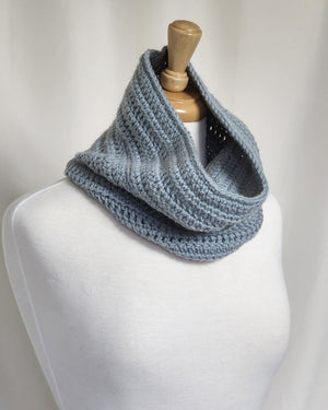 A three-quarter view of the Foggy Morning Cowl by Tanis McNally-Dawes, modeled on a white cloth mannequin.  The cowl is a steel blue, standing high on the back of the neck, swooping down in the front.
