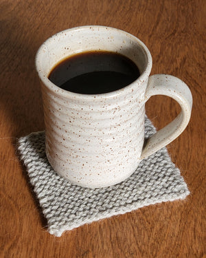 A speckled taupe ceramic mug full of dark tea sits atop a light grey coaster, knit in garter stitch.